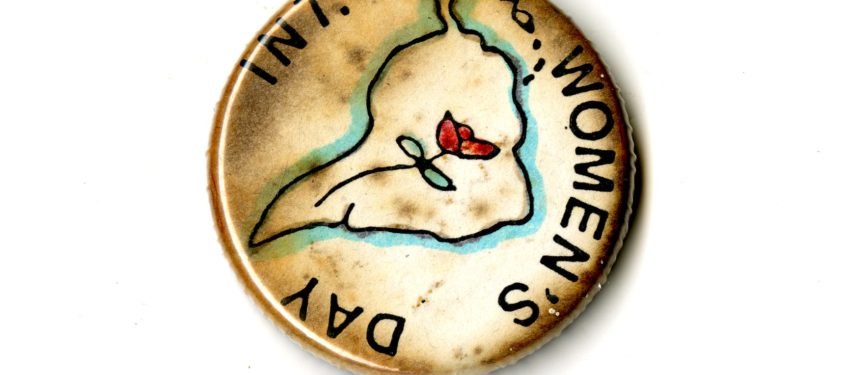 A small discolored white badge whihc the words INT. Women's Day written on it in black text. In the Centre if an outline of South America with a rose inside of it.