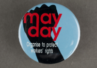 A blue badge with a black fish on it with the text May Day overlayed in red.