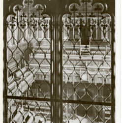 This postcard depicts a close up of two panels of a grille or iron gate that is located in the House of Commons, London. Through the cast iron you can see an empty house of parliament with wooden benches and ornate decoration.