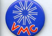 A bright blue badge with a white twelve pronged star on the top half and capital red letters on the bottom half spelling V M C.