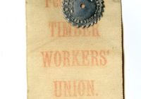 An off white, almost yellow, fabric ribbon in a long vertical rectangle with a small metal disk with spiked edged pinned to the top centre. The ribbon read, in red text, Federated Timber Workers Union.