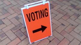 An organge a-frame board with the word 'voting' in black and a black arrow pointing right.